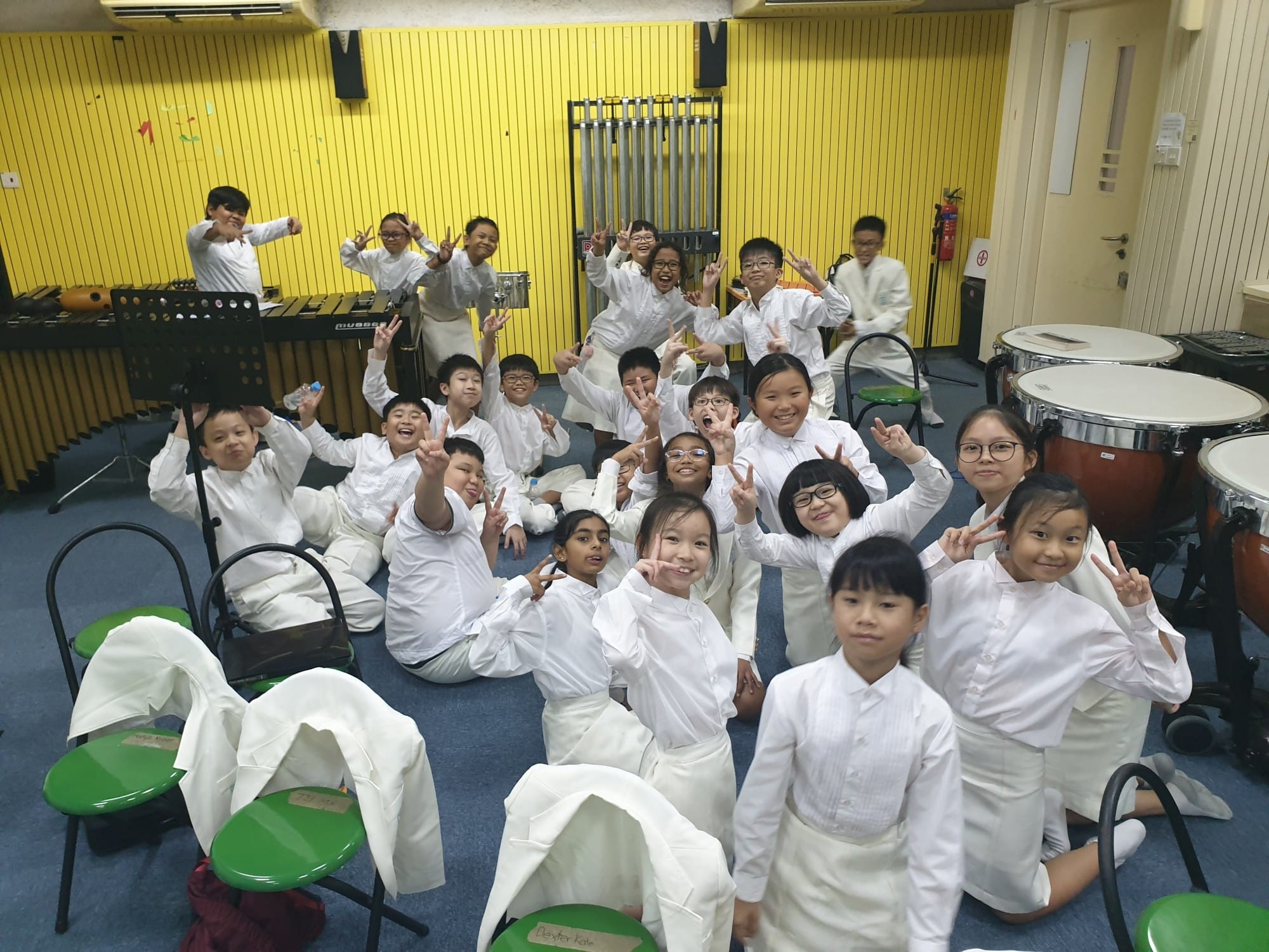 Group of children in white uniforms posing with peace signs in a music room with drums and a xylophone.